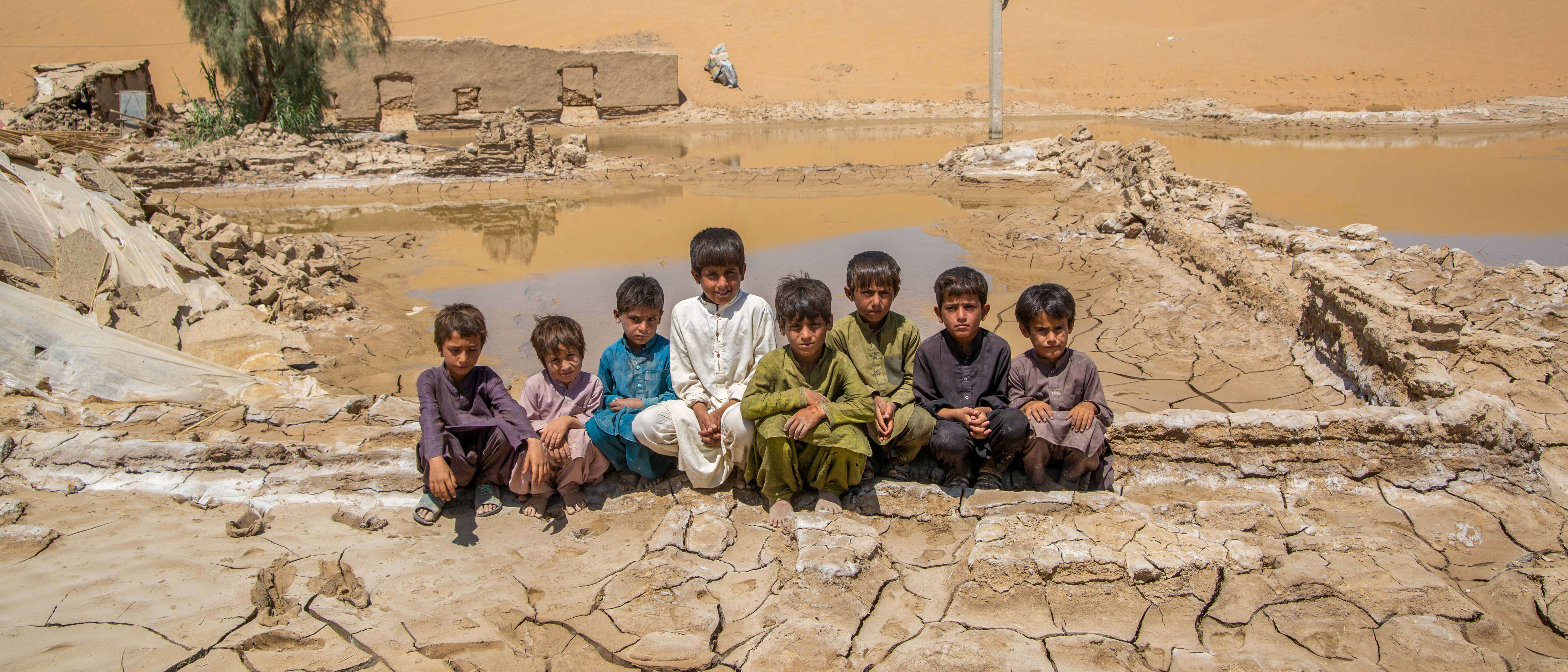 8 boys sit together outside in Pakistan, in front of a landscape devastated by flooding.